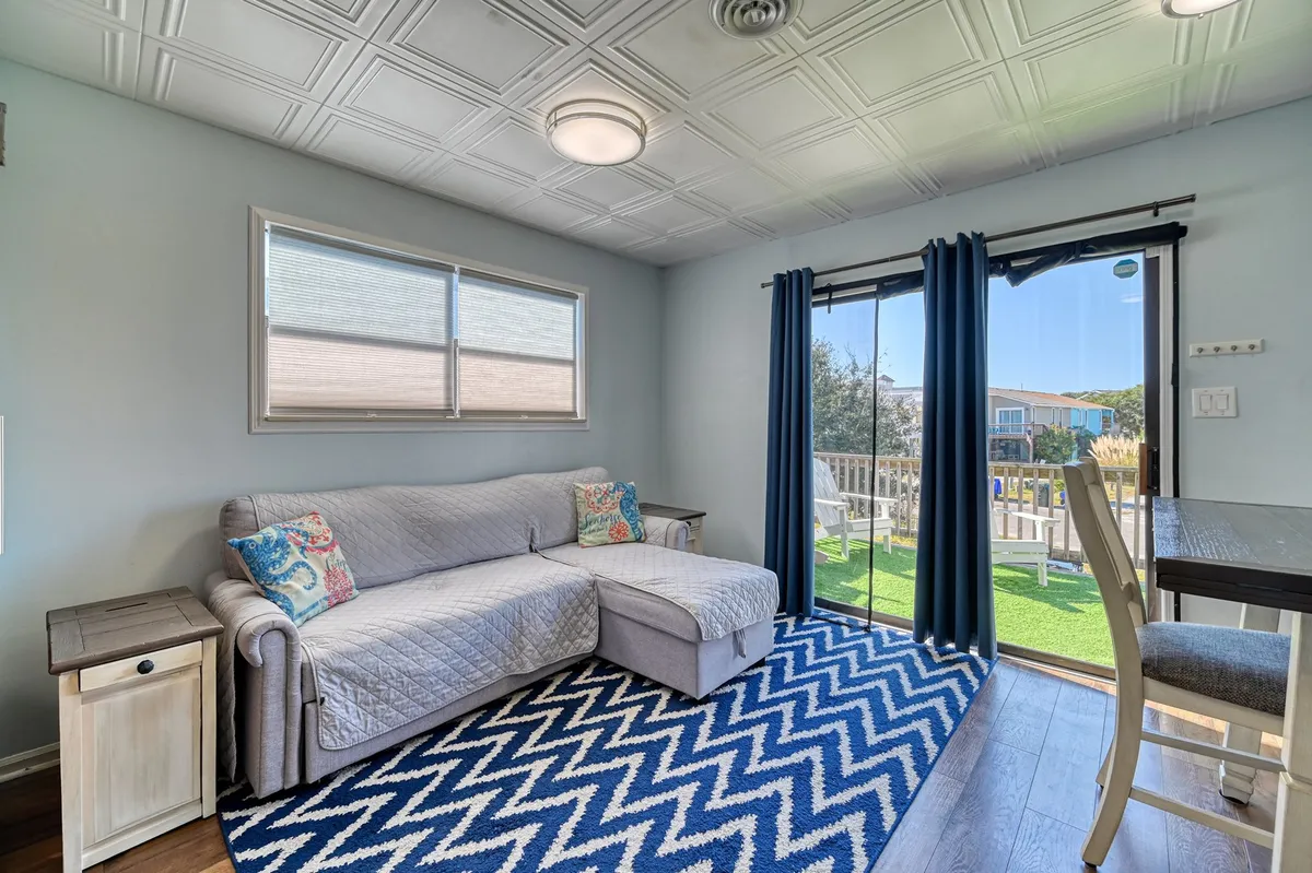 A cozy living area with a comfortable sofa, soft rug, and large sliding glass doors that flood the room with natural light and visually connect the indoors to a private sun deck. The space suggests relaxation, reading, and enjoying the changing light of morning or sunset.