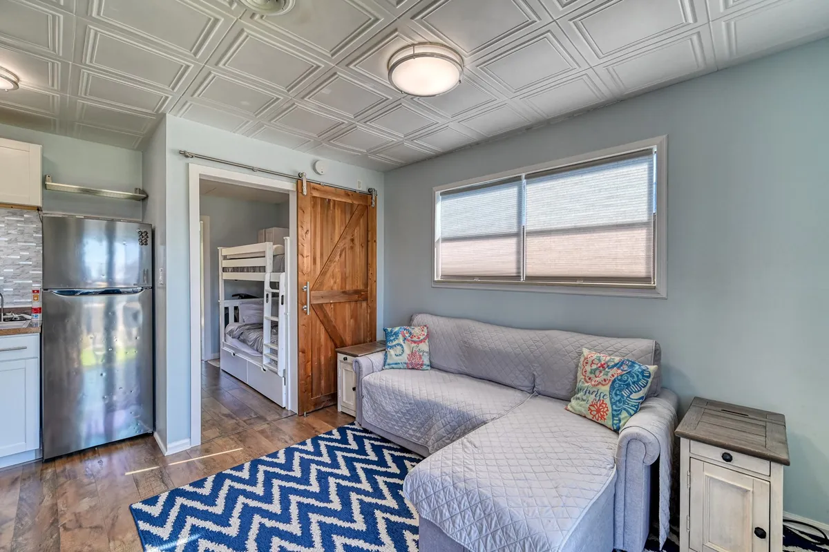 A welcoming living and dining space with a colorful-cushioned sofa, blue and white patterned rug, and bunk beds in the background. The open-plan design encourages family gatherings, play, and relaxation.