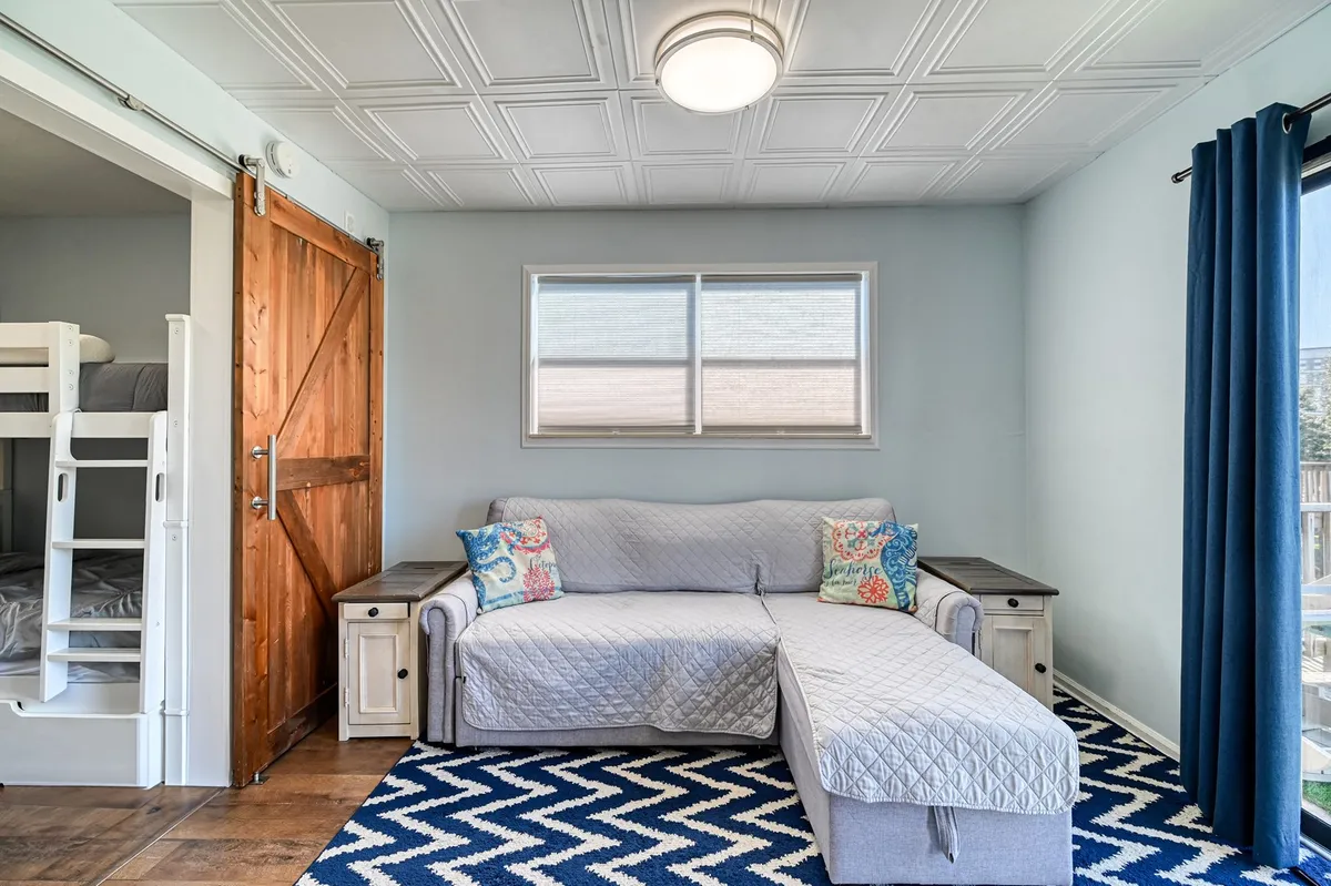 This image features a cozy living area with a sofa adorned with decorative pillows, a chevron patterned rug, and a bunk bed tucked behind a barn door. The space is filled with inviting textures and natural light, perfect for family relaxation and socializing.