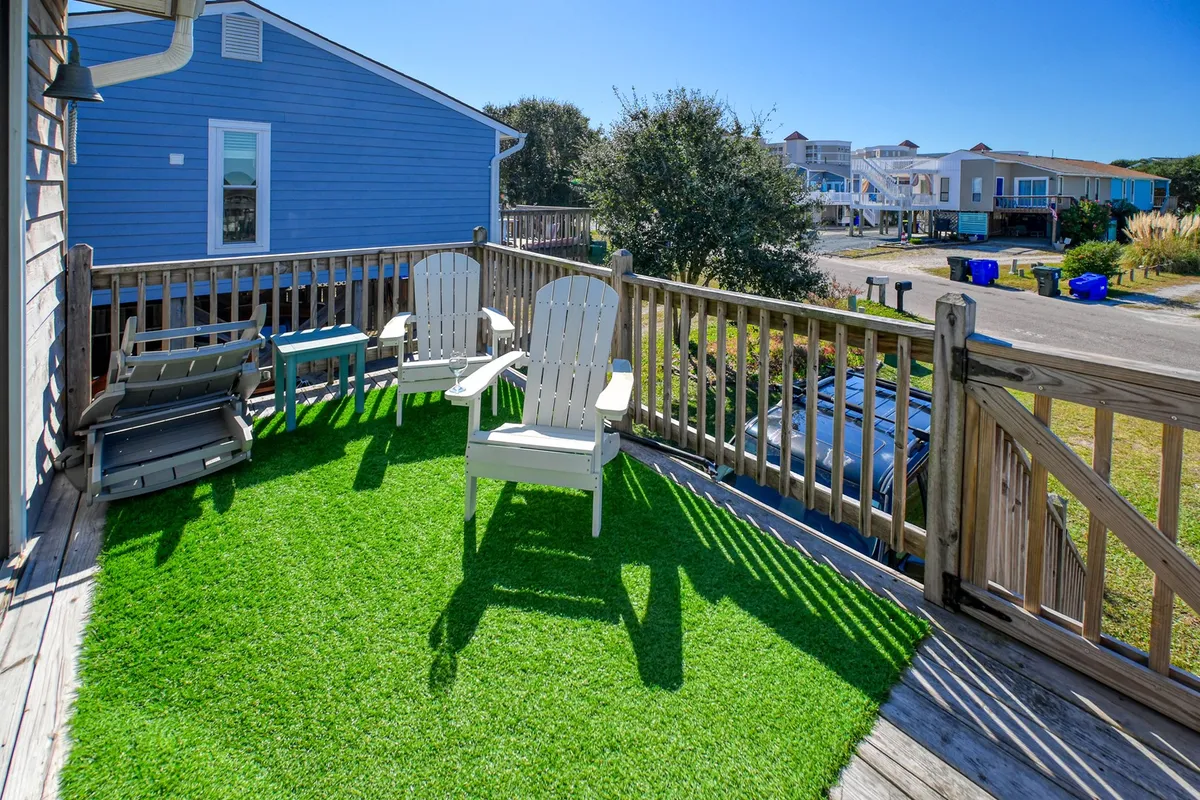 A bright outdoor deck space featuring artificial grass (Astroturf), two Adirondack chairs, a blue side table, and a wooden railing. Sunlight bathes the area, and the setting feels private and inviting—perfect for relaxing or socializing.