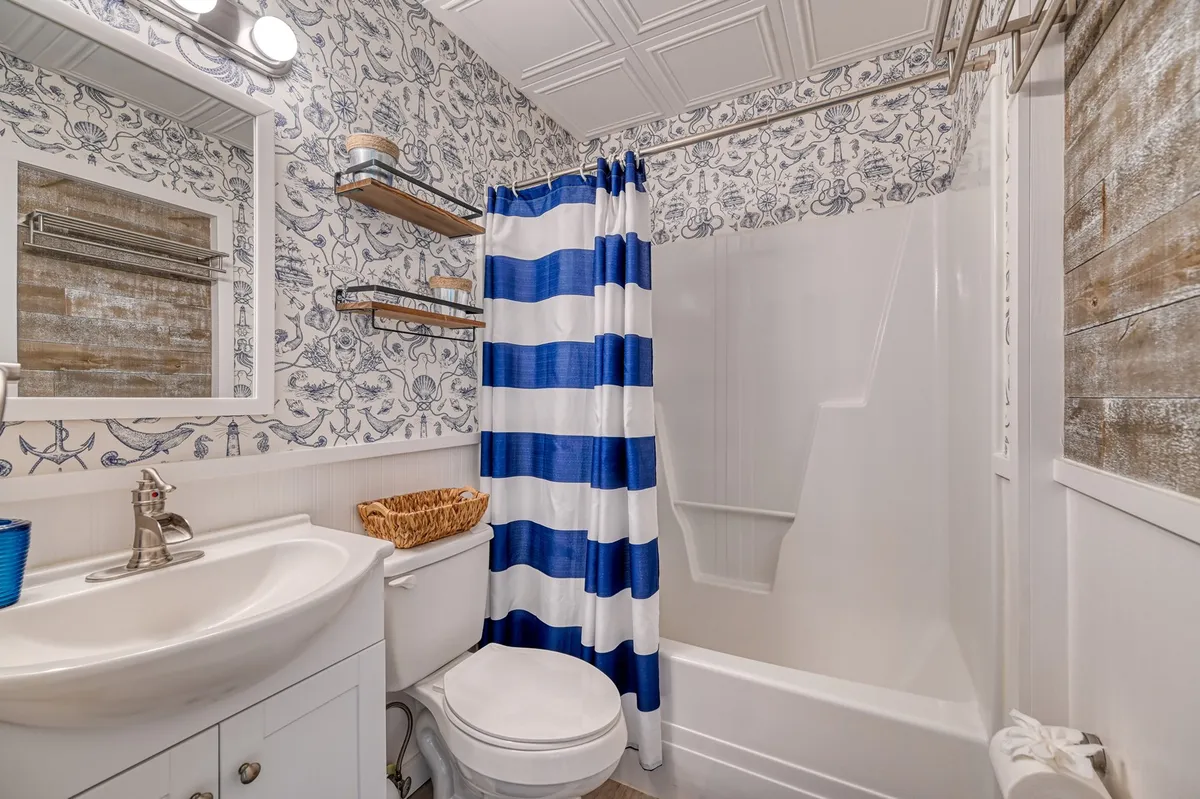A bright and inviting bathroom features a cheerful striped shower curtain, ornate wallpaper, and a modern vanity. The space looks fresh and welcoming, perfect for rinsing off after a day at the beach or getting the little ones ready for bed.