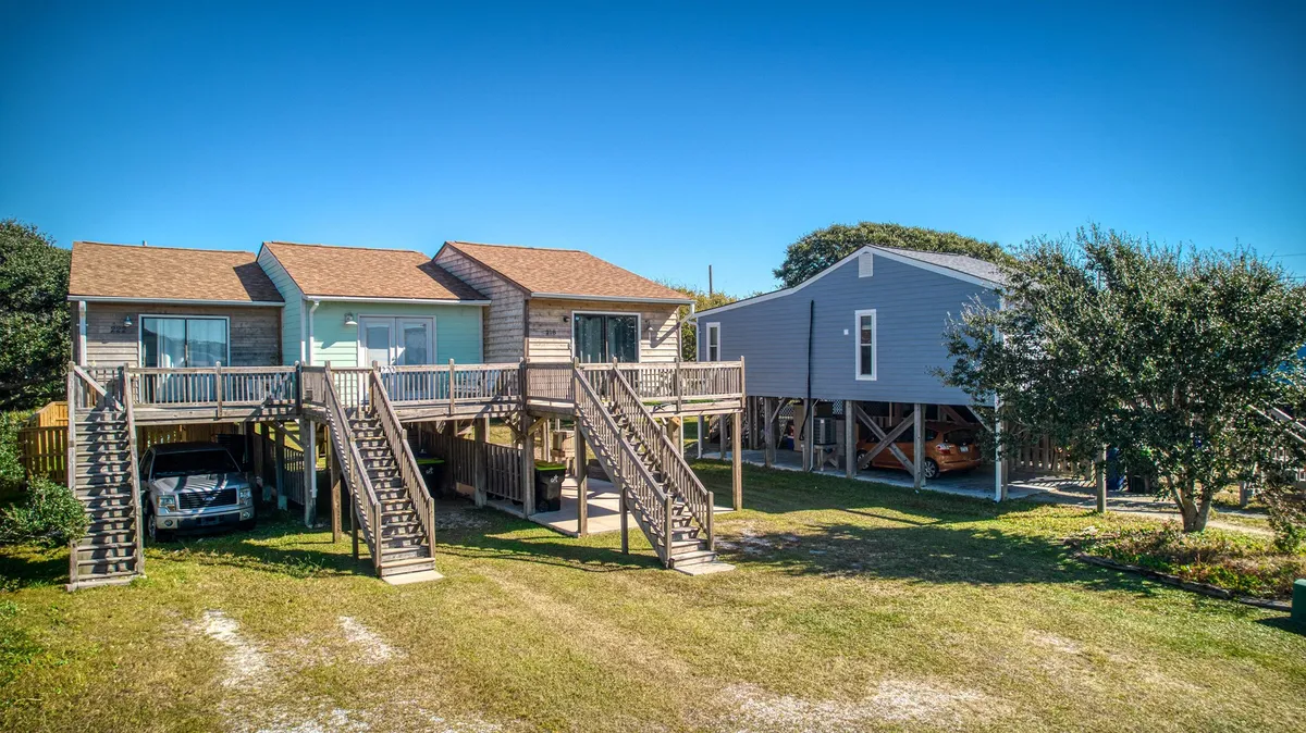 Two elevated seaside houses with stairs leading up and lush greenery surrounding them, conveying a welcoming, family-friendly neighborhood vibe. The image suggests space for gatherings, outdoor play, and easy access to both beach and sound.