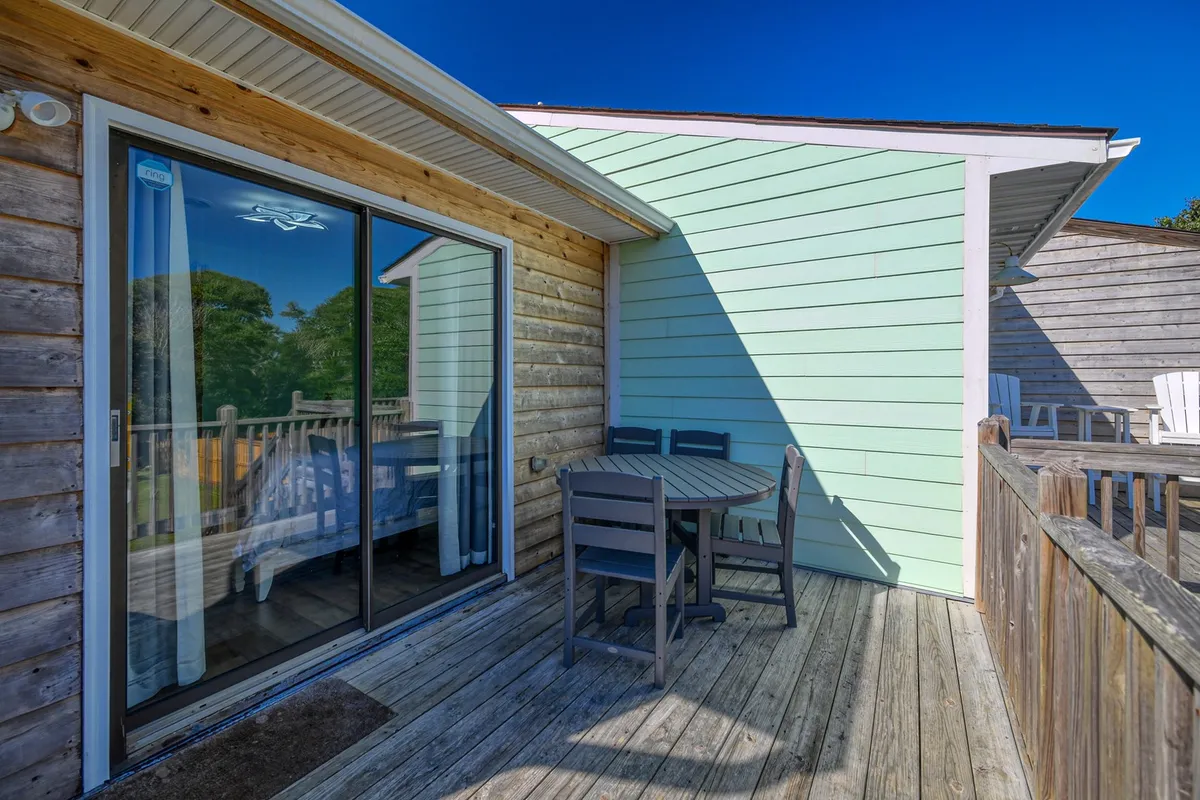 A welcoming exterior view featuring a sliding glass door, outdoor dining table, and deck railing. The space is ideal for dining al fresco, relaxing, or gathering with friends and family—activities easily shared with a pet.
