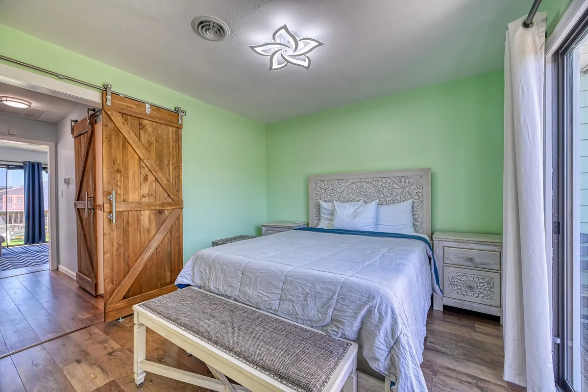 An intimate view of a bedroom featuring a comfortable bed, a stylish decorative light fixture reminiscent of a chandelier fan, and a rustic wooden barn door. The space is bright with natural light, emphasizing calm and comfort.
